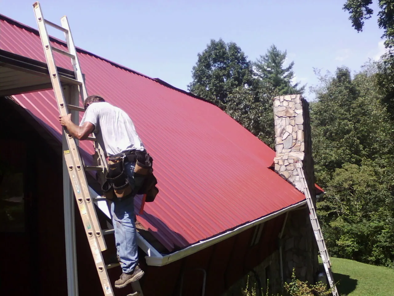 Roofer on a ladder beside a red metal roof and stone chimney — Soffit & Fascia Repair by Roofing Brothers in Friendsville